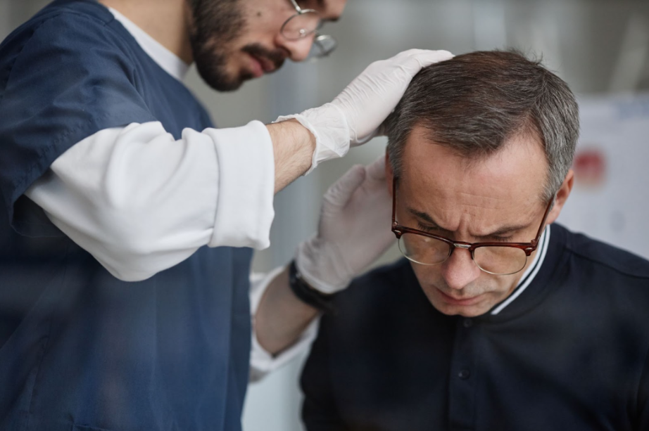 A medical professional wearing blue scrubs and white gloves carefully examines the scalp and thinning hair of a man with glasses who is seated and looking downward.