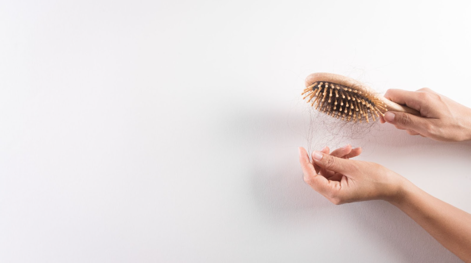 A person's hands holding a wooden hairbrush filled with tangled strands of fallen brown hair against a plain white background.